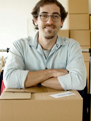 Smiling man with eyeglasses standing behind a cardboard box in a startup office setting.